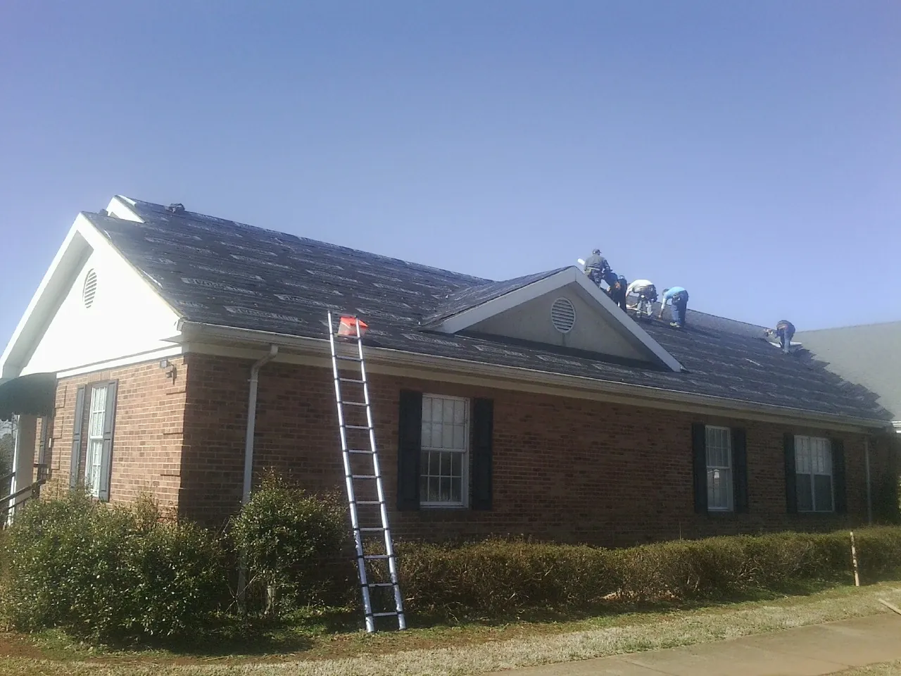 Crew tearing shingles off a brick ranch roof with a ladder set — Metal Roof Repair work by our crew in Gwinner