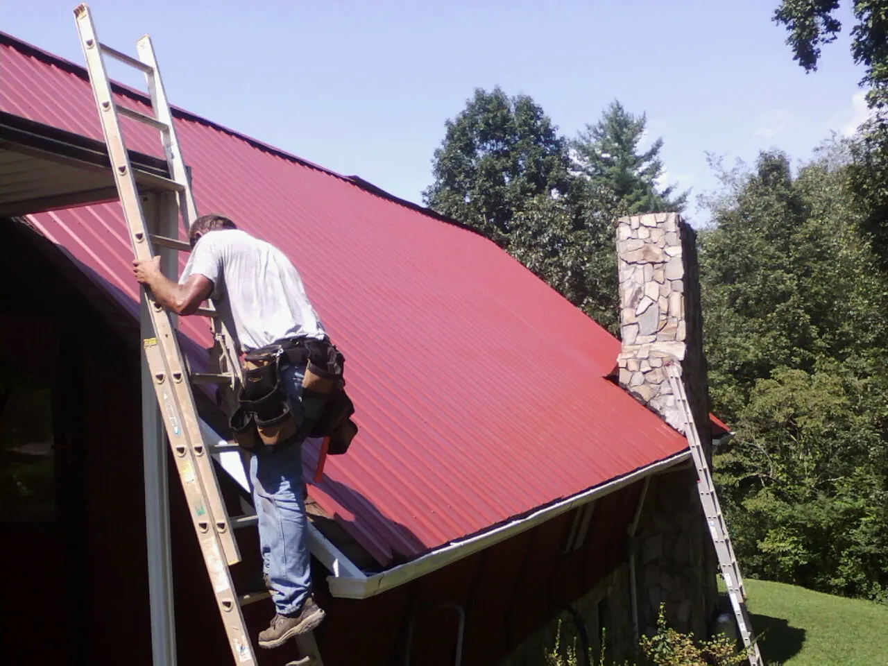 Roofer on a ladder beside a red metal roof and stone chimney — Soffit & Fascia Repair by Roofing Brothers in Gwinner